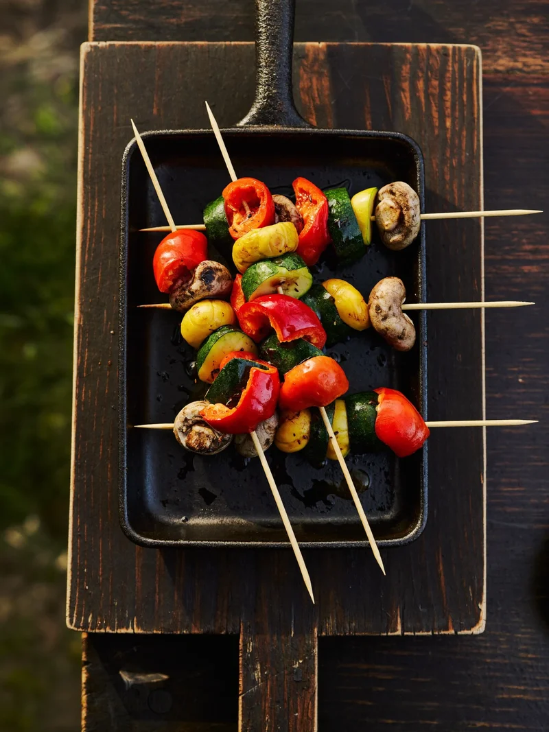 Illustration: Gussgrillpfanne mit Gemüsespiessen auf rustikalem Holzbrett