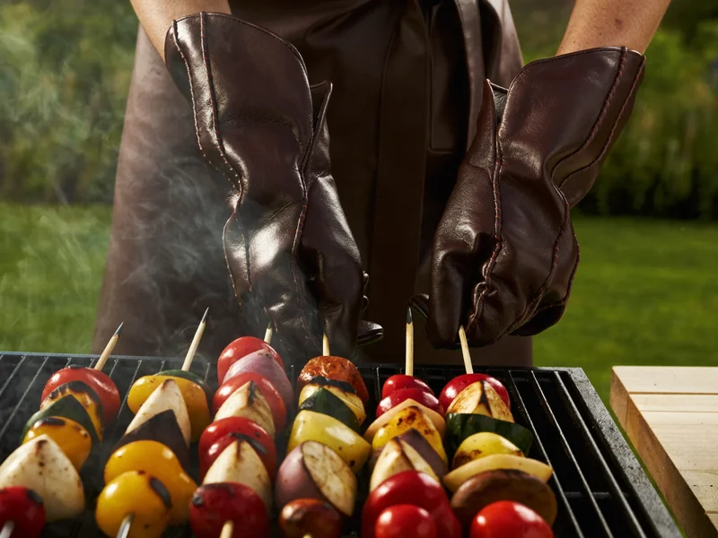 Illustration: behandschuhte Hände wenden Gemüsespiesse über dem Grillrost